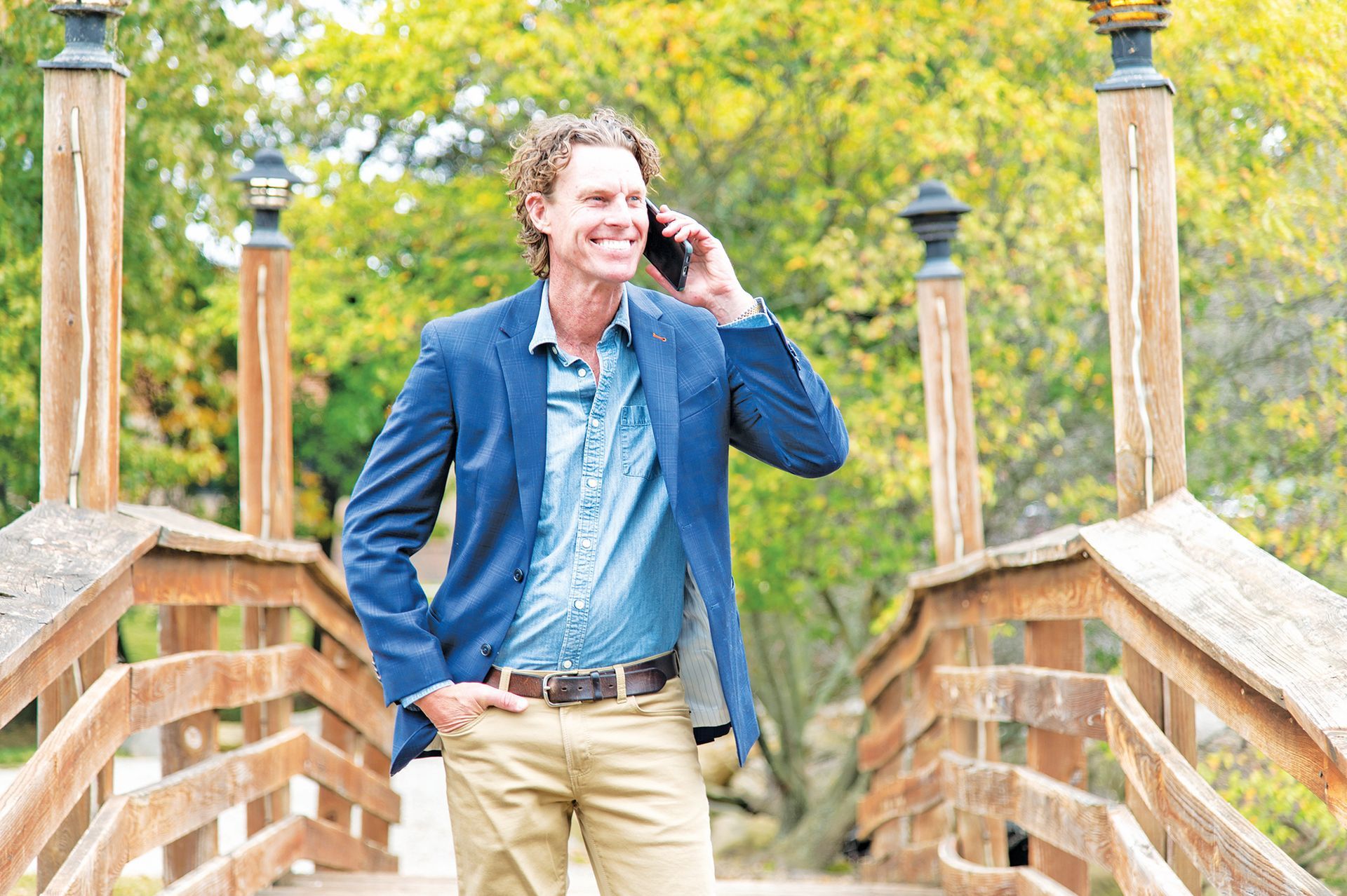 A smiling person in a blue blazer and khakis stands on a wooden bridge outdoors, talking on a mobile phone.
