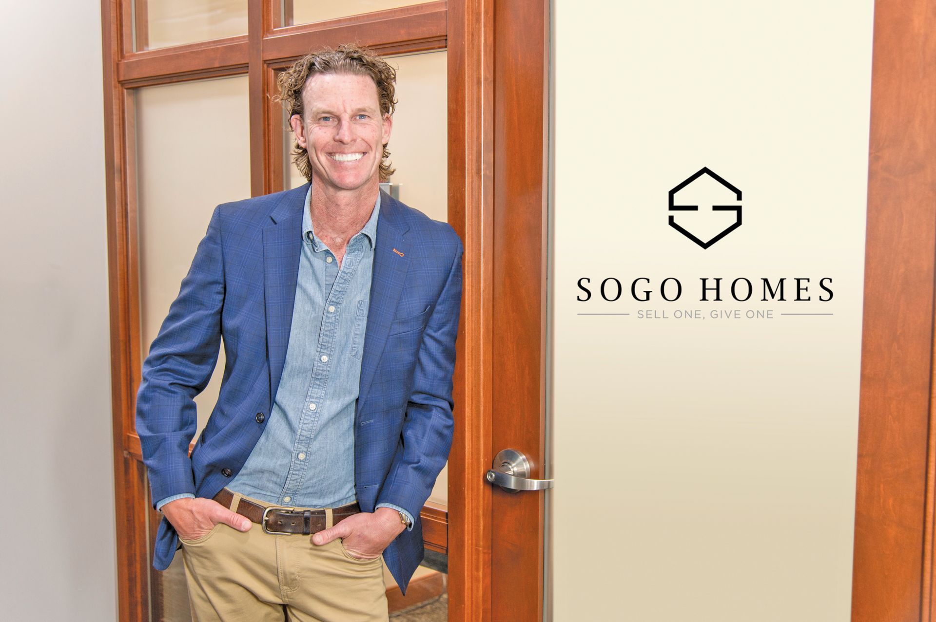 A smiling person wearing a blue blazer and khakis stands in a doorway next to a sign for SOGO HOMES.