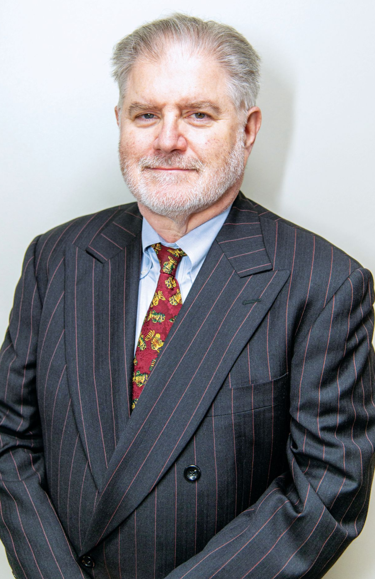A man wearing a grey pinstripe suit, light blue dress shirt, and a red patterned tie standing against a white background.
