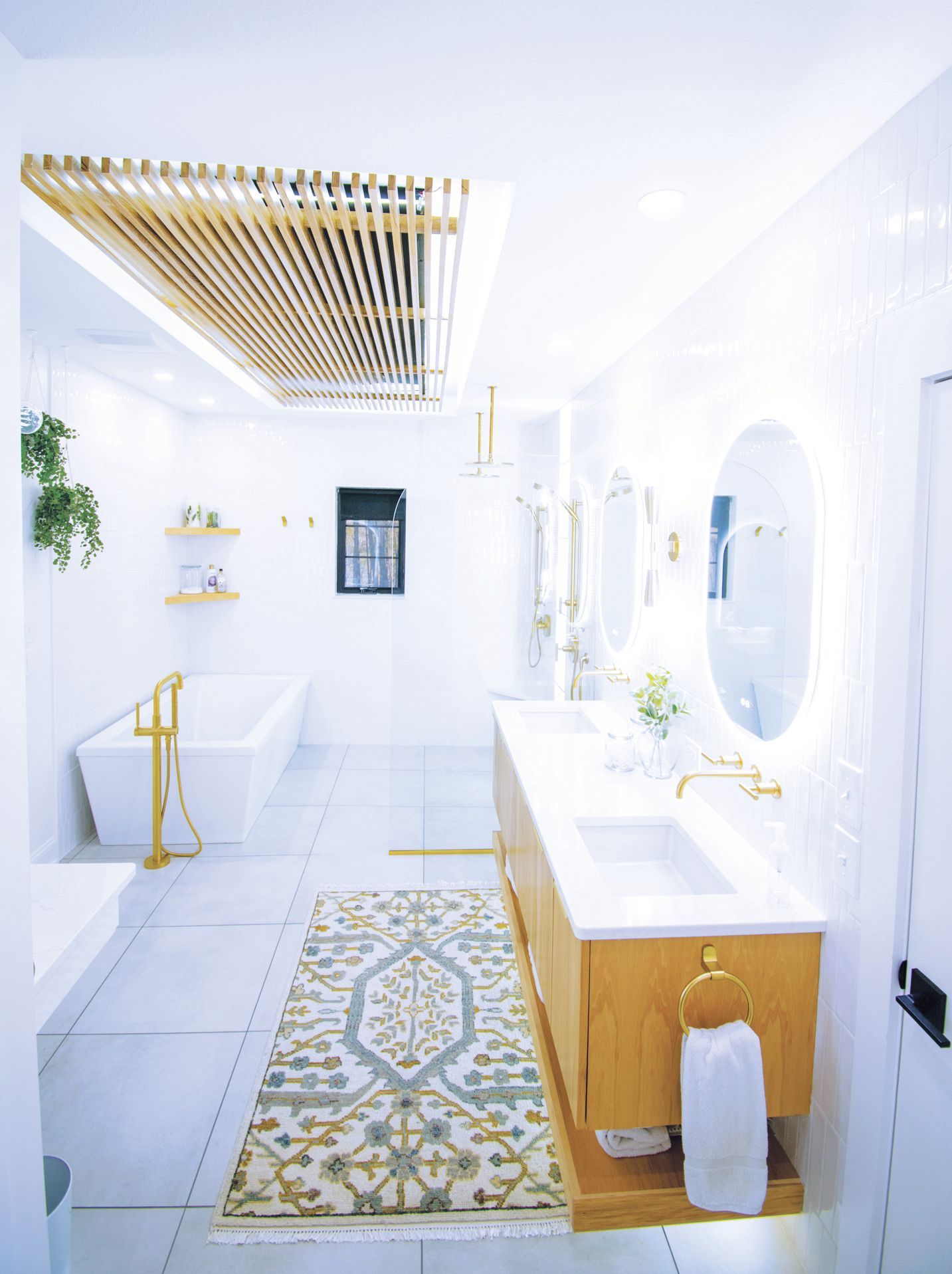 Modern bathroom with white walls, a freestanding tub, wood-toned vanity, gold fixtures, and an intricate patterned rug.