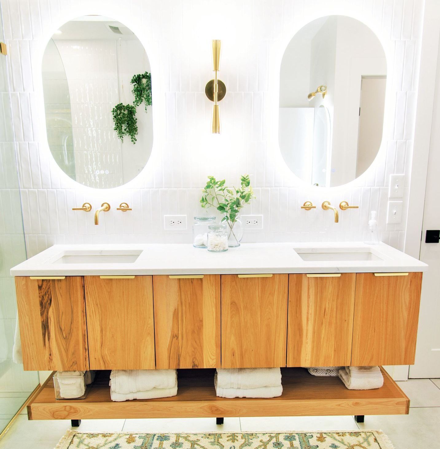 A modern double-sink wooden vanity with white countertops, gold fixtures, two oval backlit mirrors, and open shelving.
