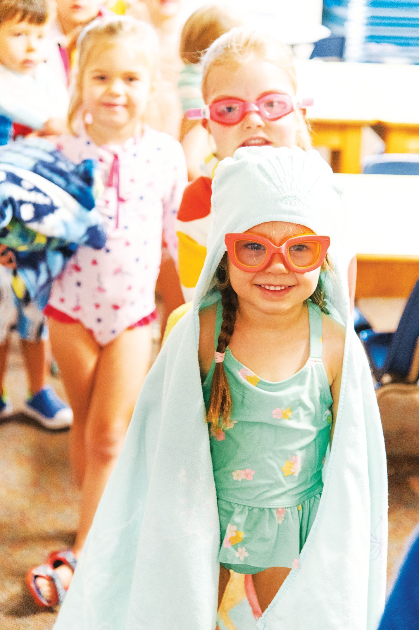 Children in swimsuits and goggles stand in a row; a girl in the foreground wears a light blue hooded towel.