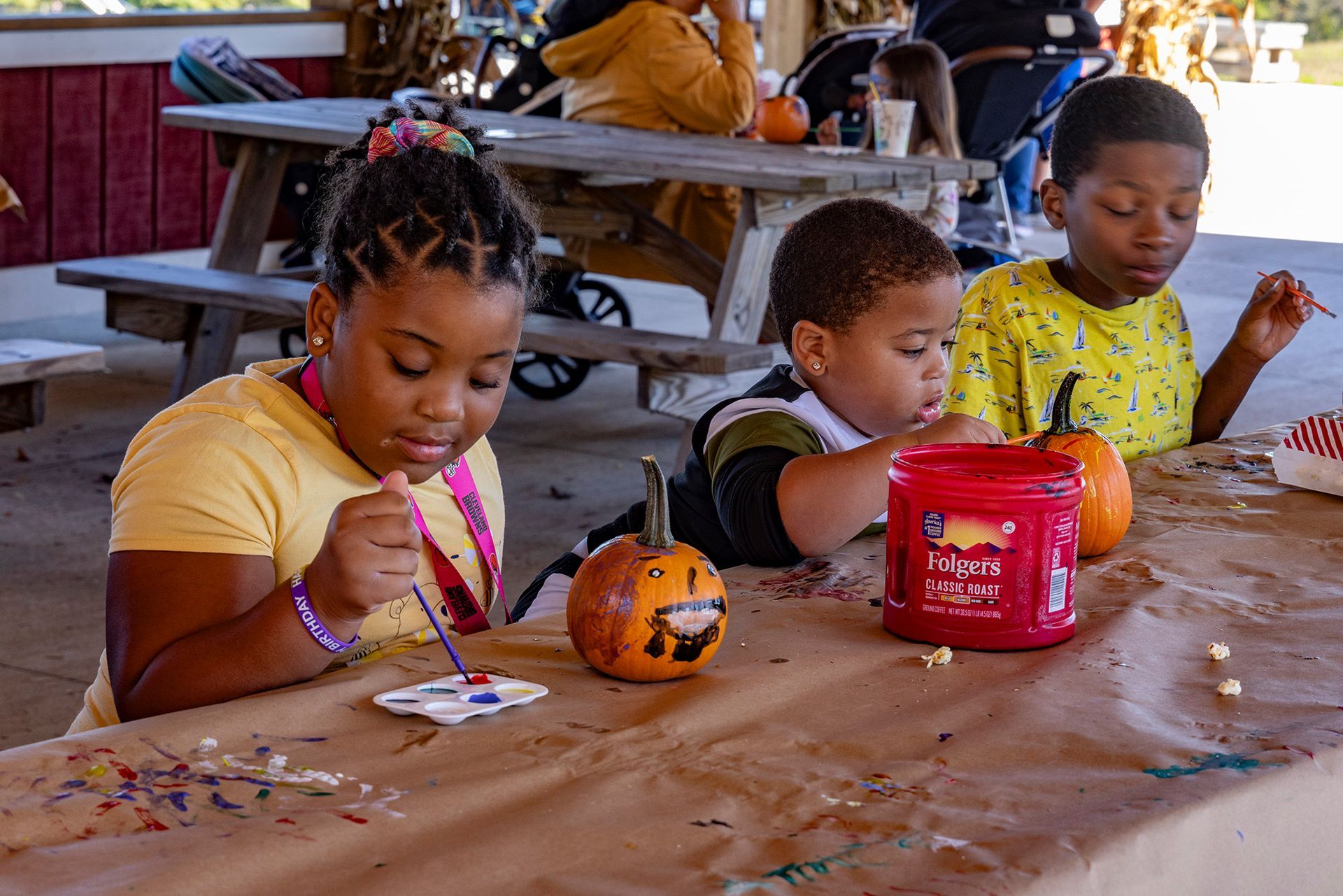 Children paint small pumpkins at a wooden table in an outdoor, covered area.