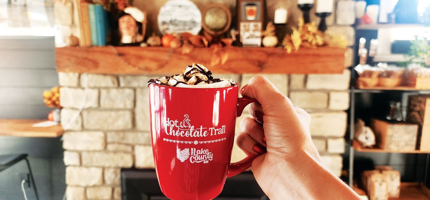 A hand holds a bright red mug filled with hot cocoa and whipped cream in front of a cozy stone fireplace.