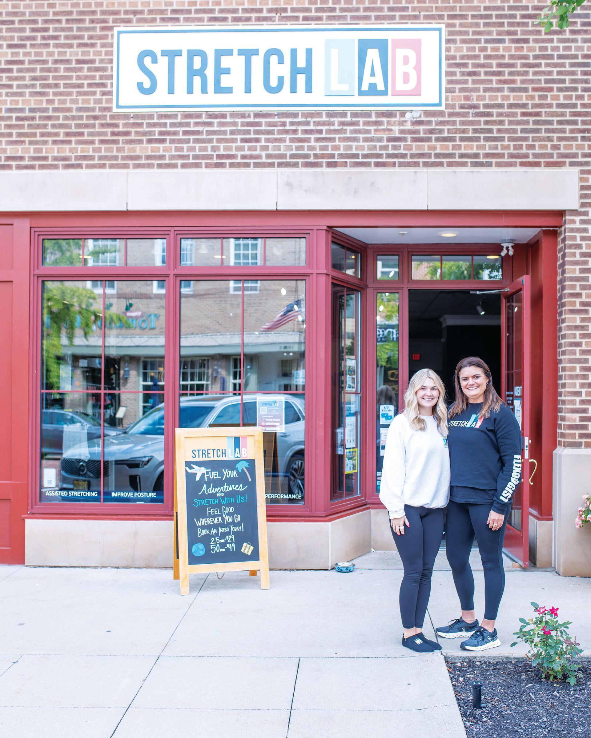 Two people stand smiling in front of a brick building with a StretchLab sign and an A-frame chalkboard sign.