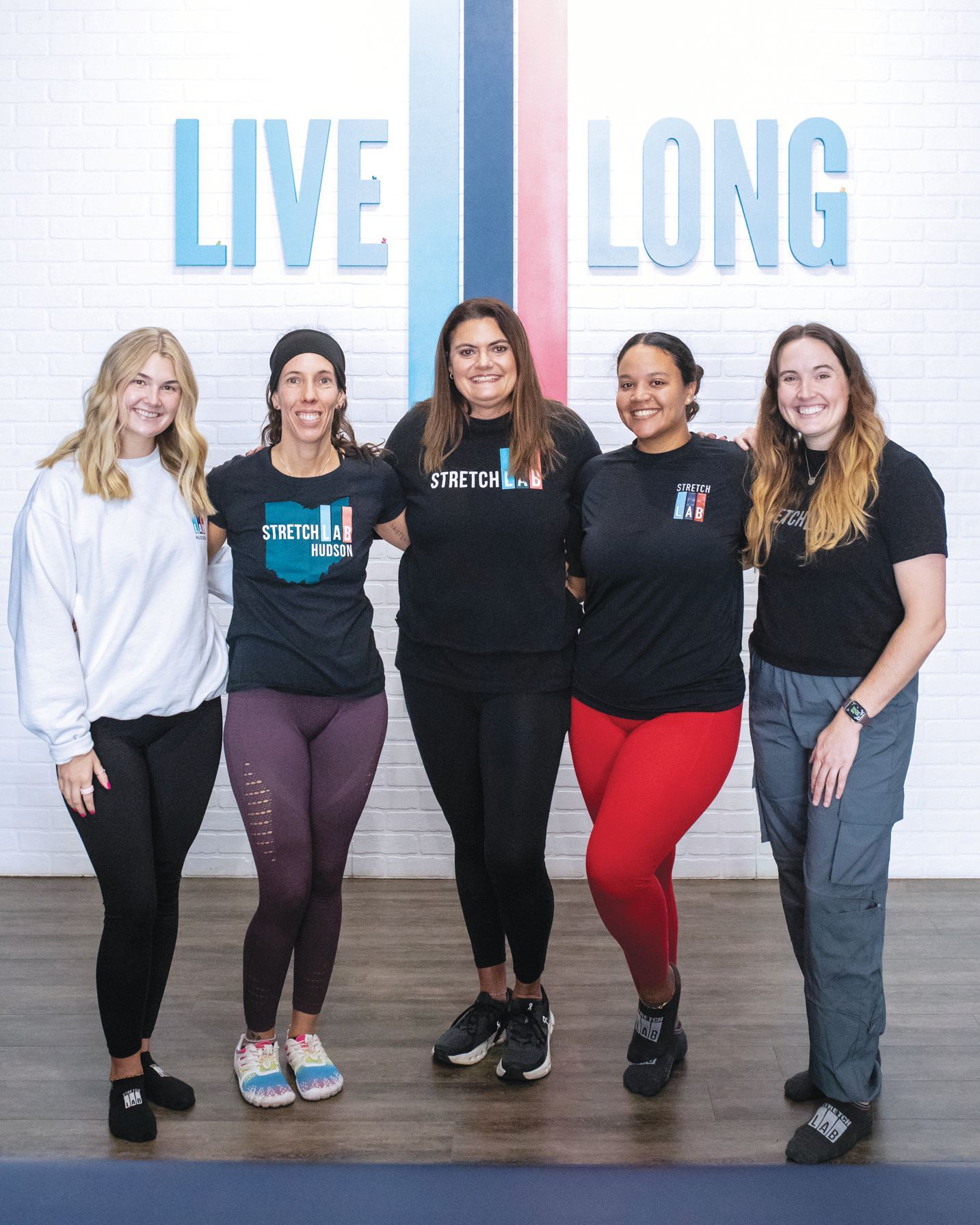 Five smiling people stand in a line against a white brick wall with the words 