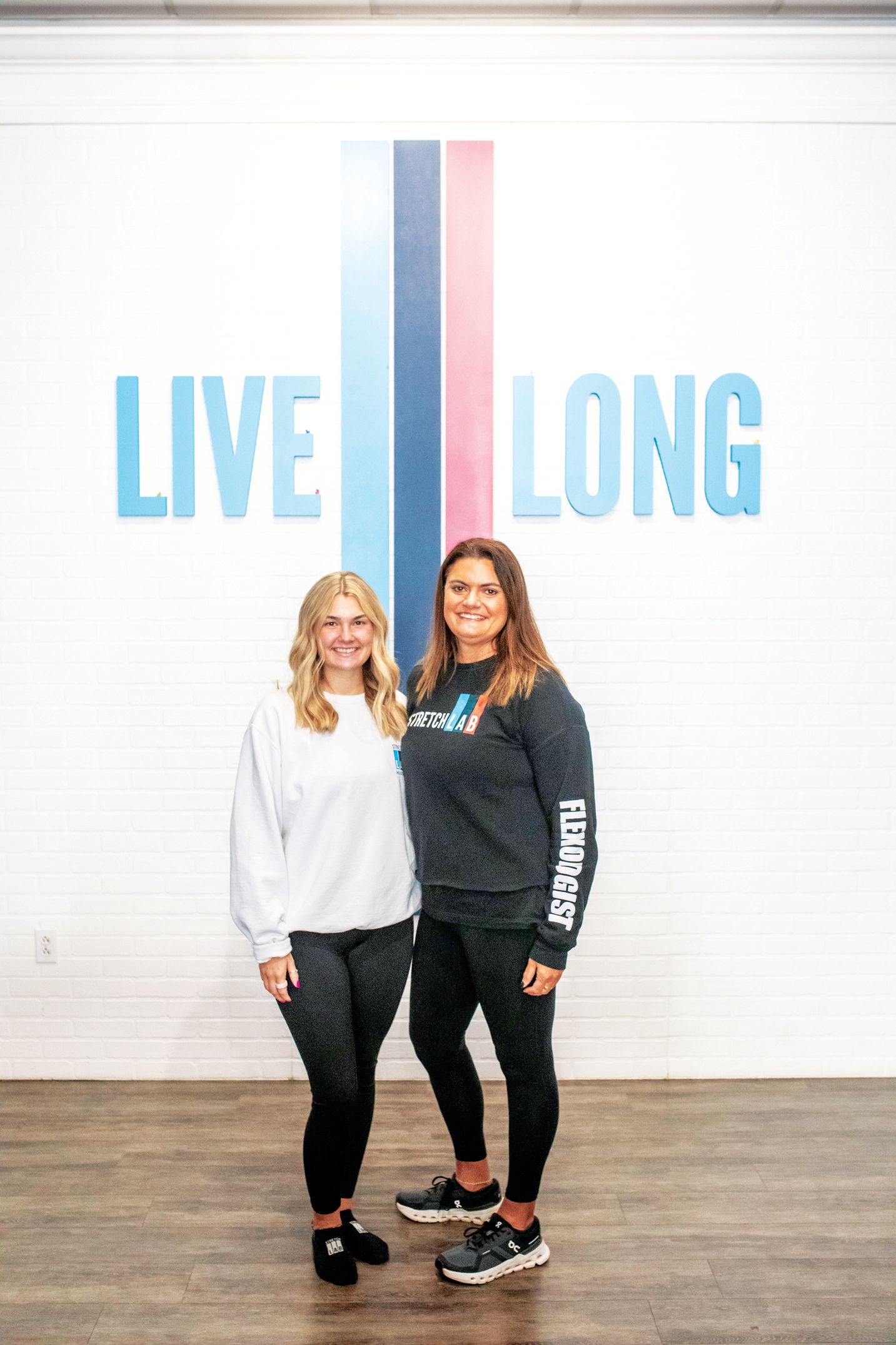 Two people stand in front of a white wall with the blue text 