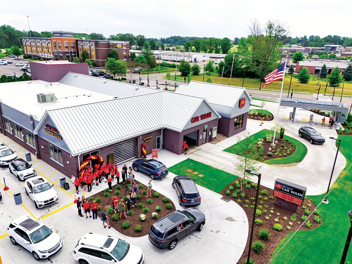 An aerial view shows a new commercial building with cars, a crowd of people in red shirts, and an American flag.