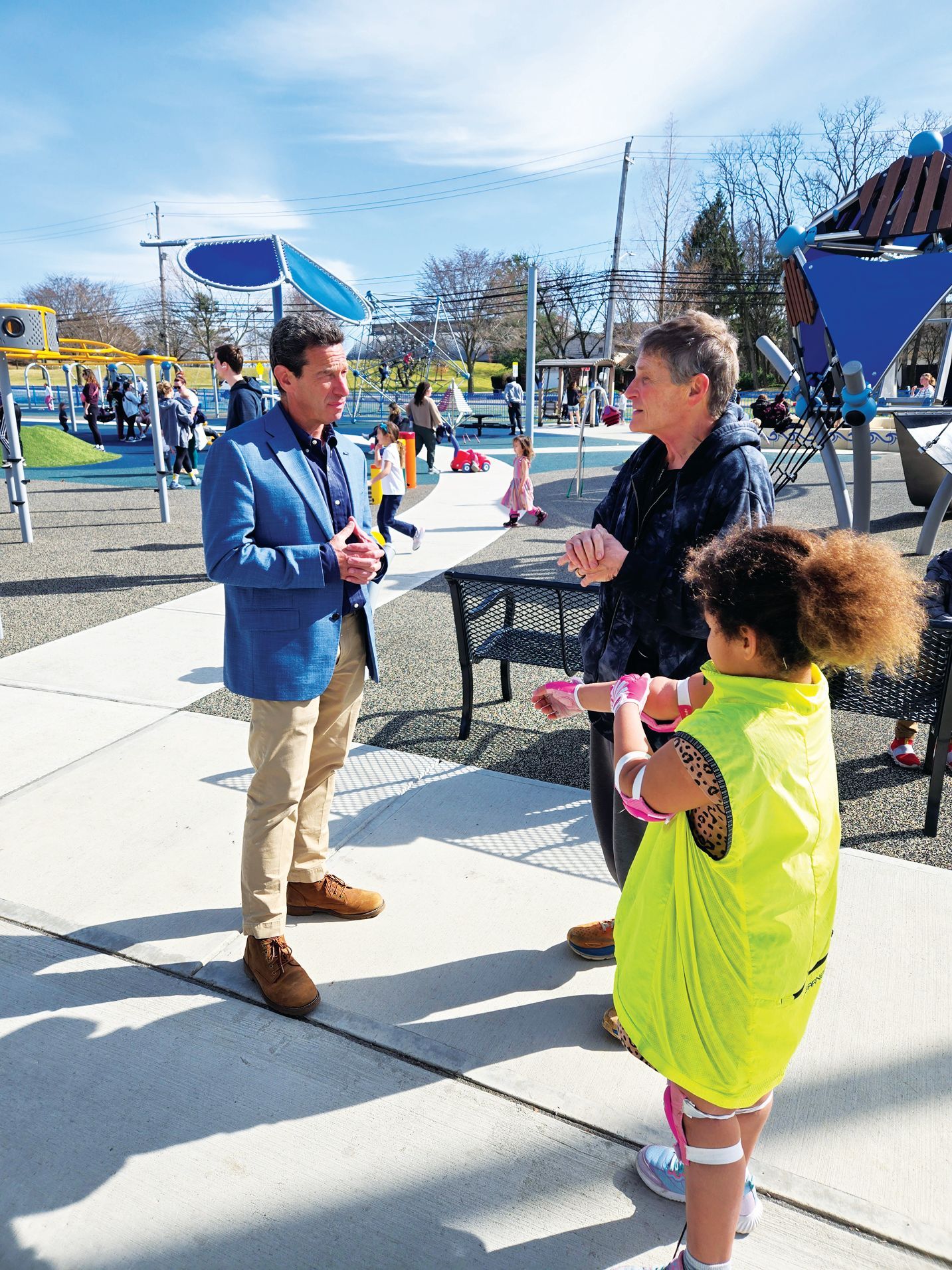 A person in a blue blazer speaks with two others at a brightly colored outdoor playground on a sunny day.