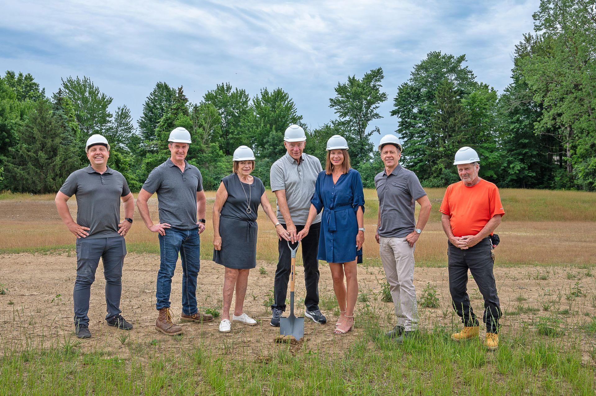 Seven people in hard hats stand in a field for a groundbreaking ceremony, with a shovel positioned in the dirt.
