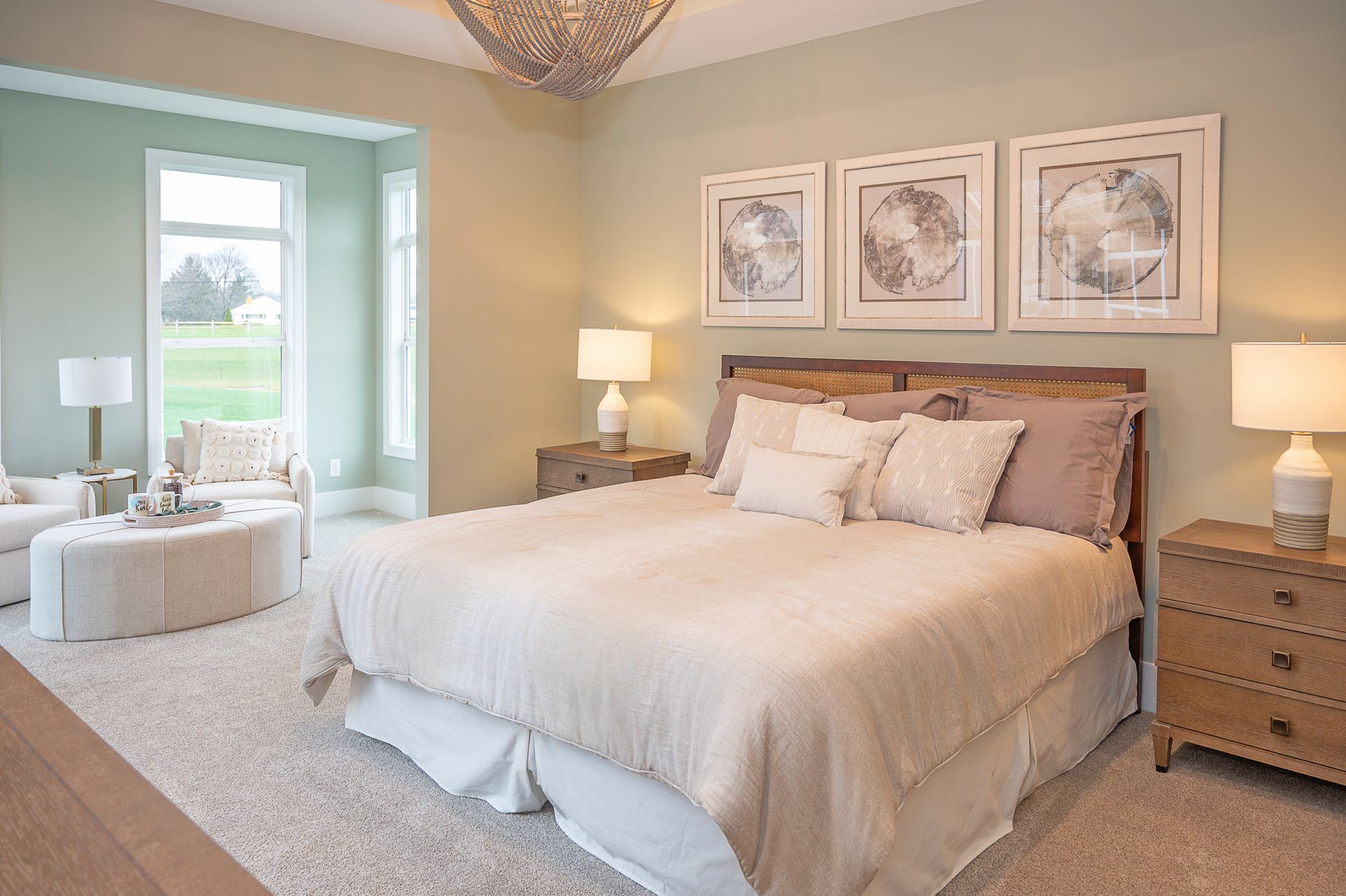A bright, neutral-toned bedroom with a large bed, three framed wall prints, wooden nightstands, and a sitting area.