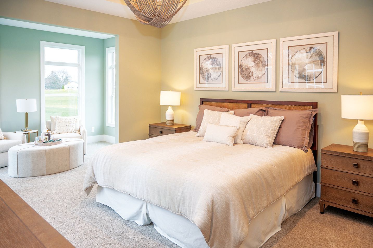 A bright, neutral-toned bedroom with a large bed, two nightstands with lamps, a circular ottoman, and three wall prints.