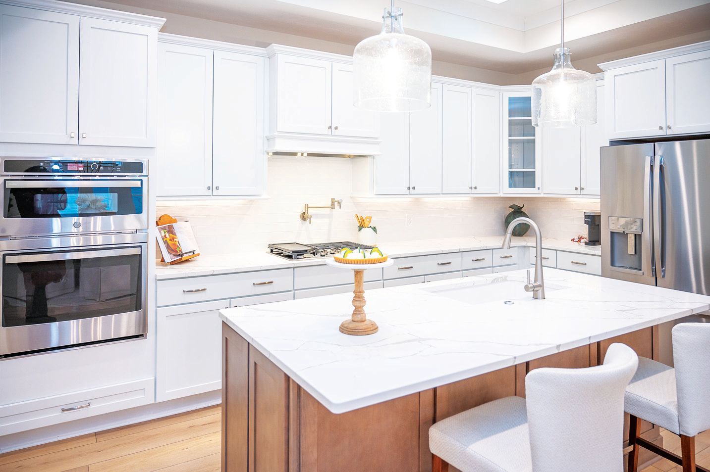 Modern white kitchen featuring a large island with two chairs, stainless steel appliances, and a wood-toned base cabinet.