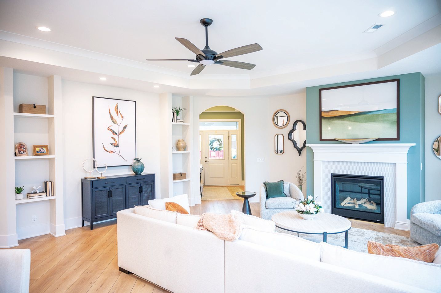 A bright living room with a white sofa, dark sideboard, fireplace, teal accent wall, and ceiling fan over hardwood floors.