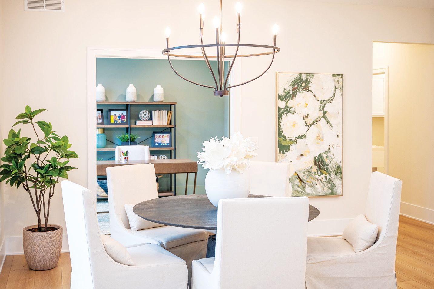 A circular wooden dining table with four white upholstered chairs sits under a chandelier, with a green room in the back.