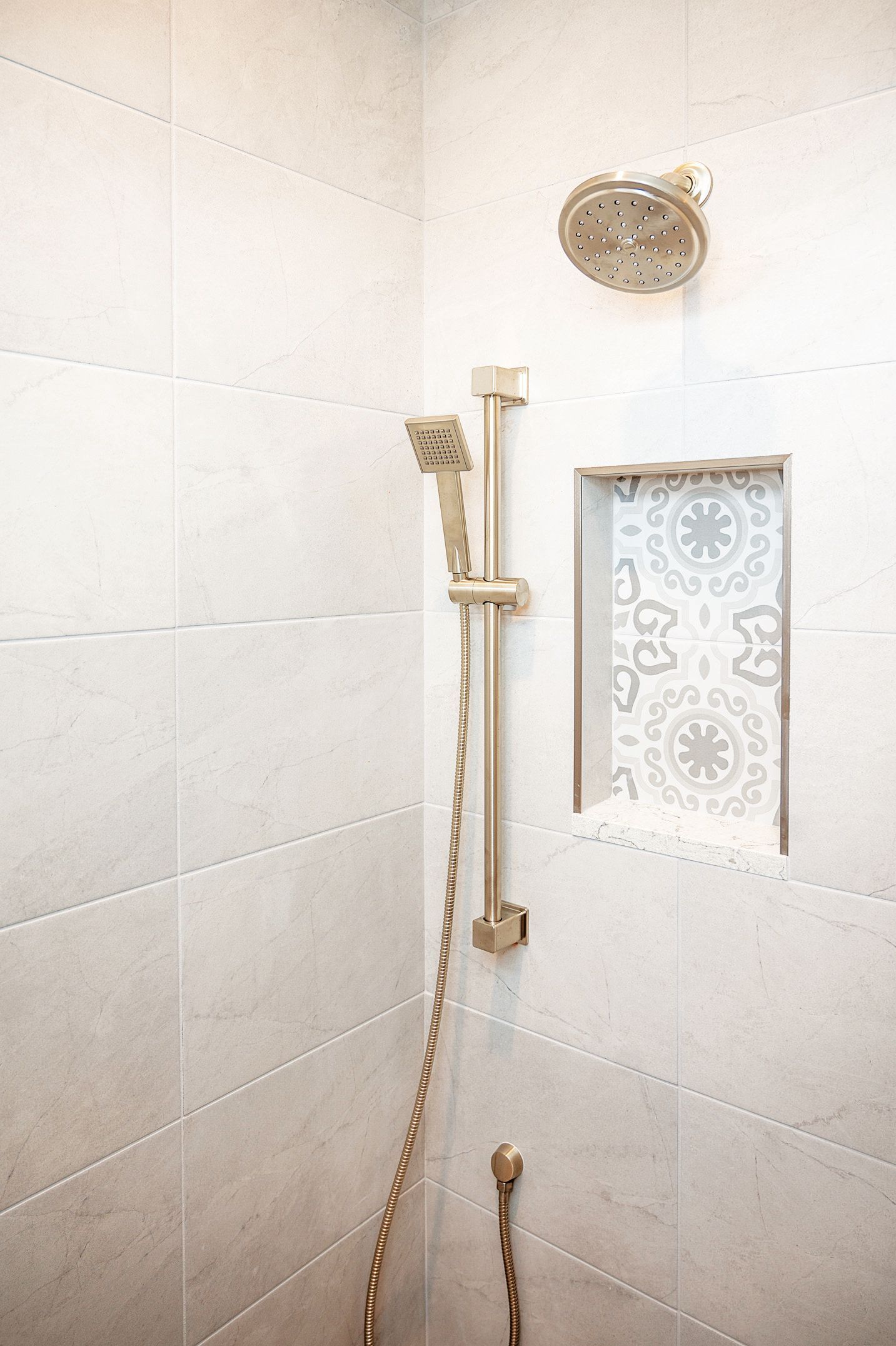 A shower wall with light neutral tiles, a gold shower head, a handheld sprayer on a bar, and a decorative tiled niche.