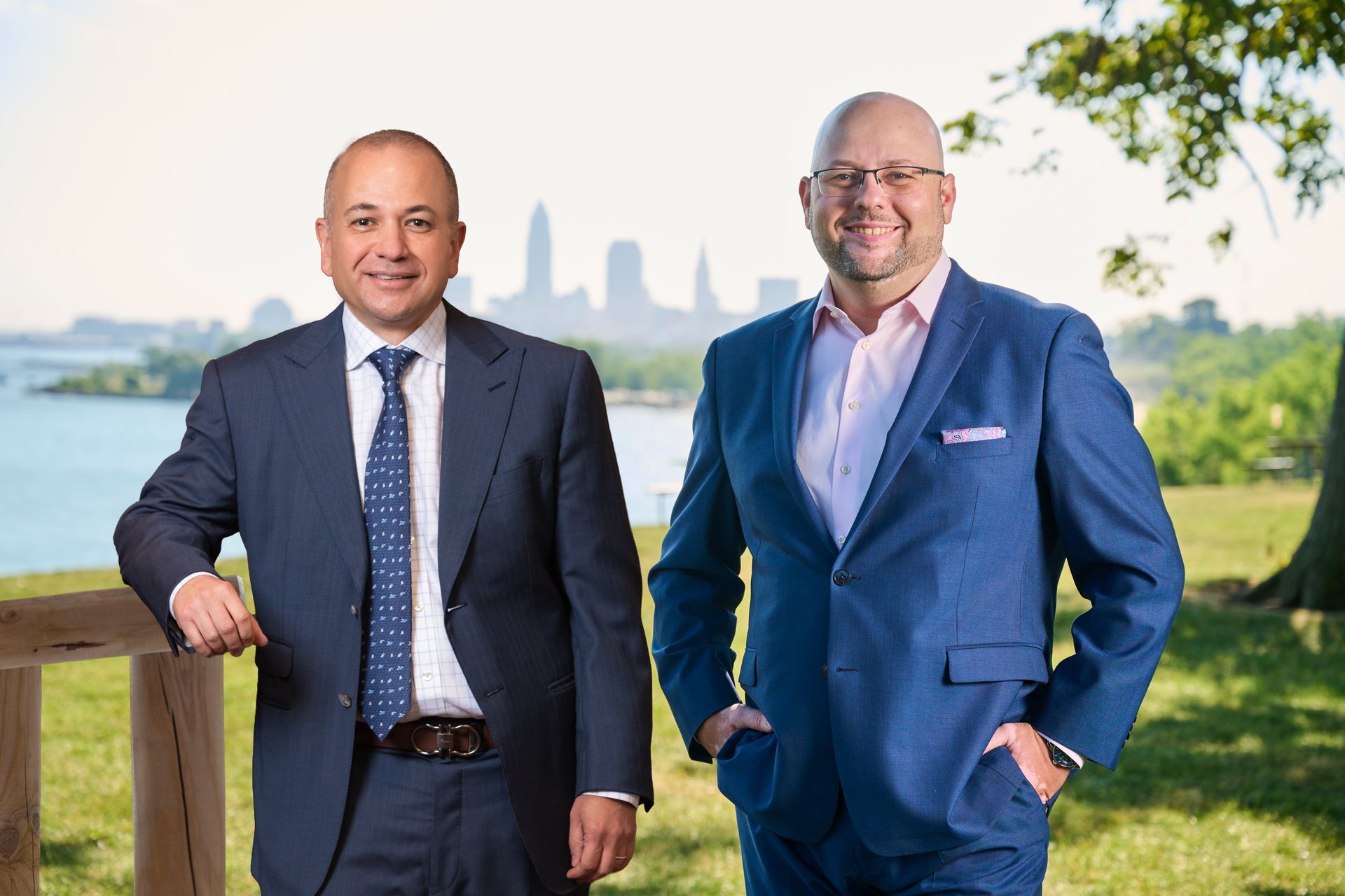 Two men in suits standing outdoors near a wooden railing with a city skyline and body of water in the background.