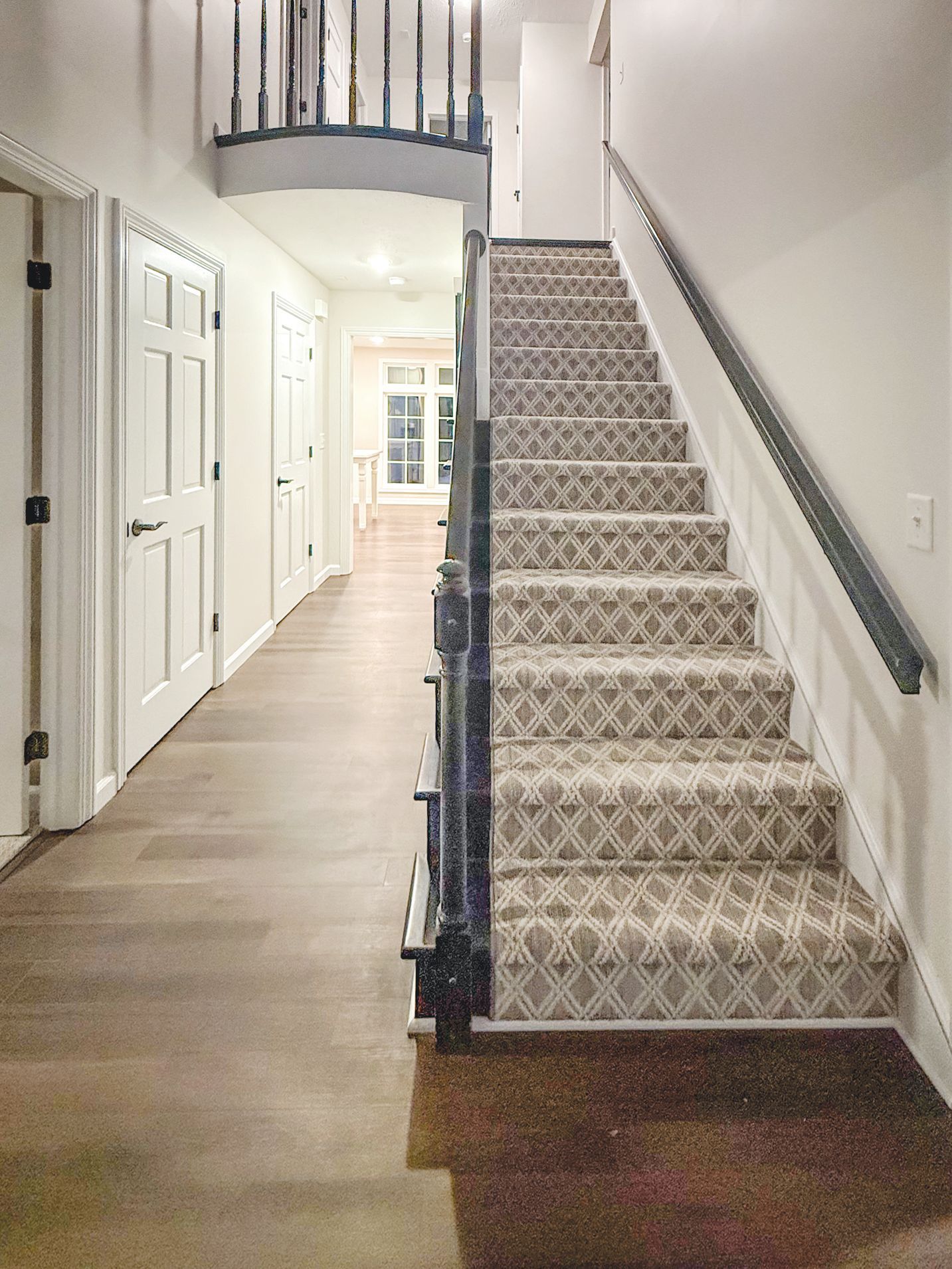 A carpeted staircase with a dark railing sits beside a hardwood hallway leading to a bright, windowed room.