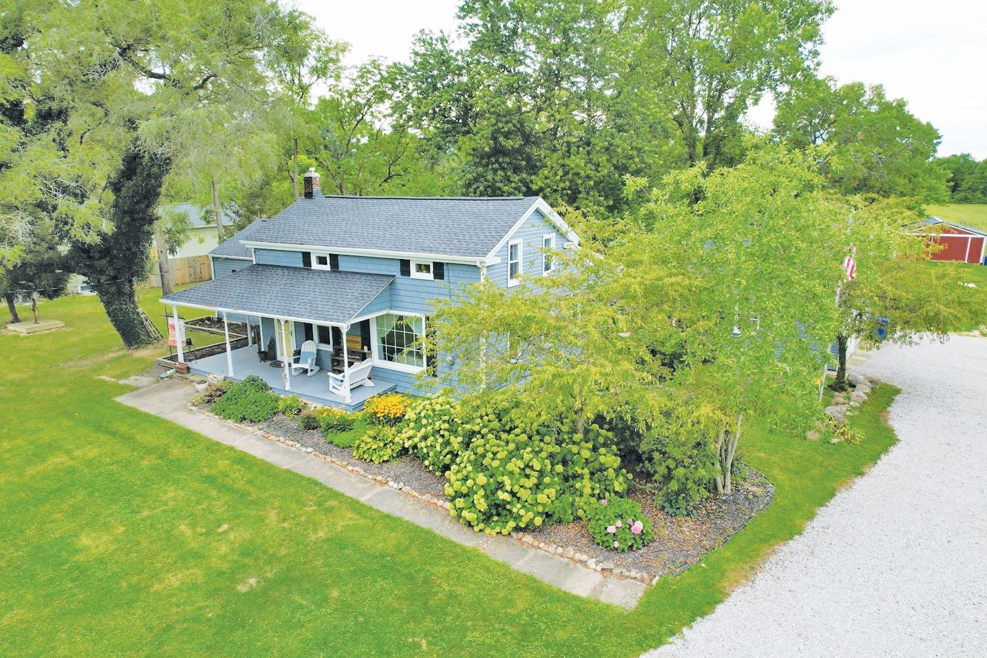 An aerial view of a blue, two-story house with a wraparound porch, surrounded by lush green trees and a gravel driveway.