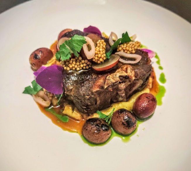 A gourmet plate featuring a seared lamb chop, roasted potatoes, pickled mustard seeds, and edible purple flowers.
