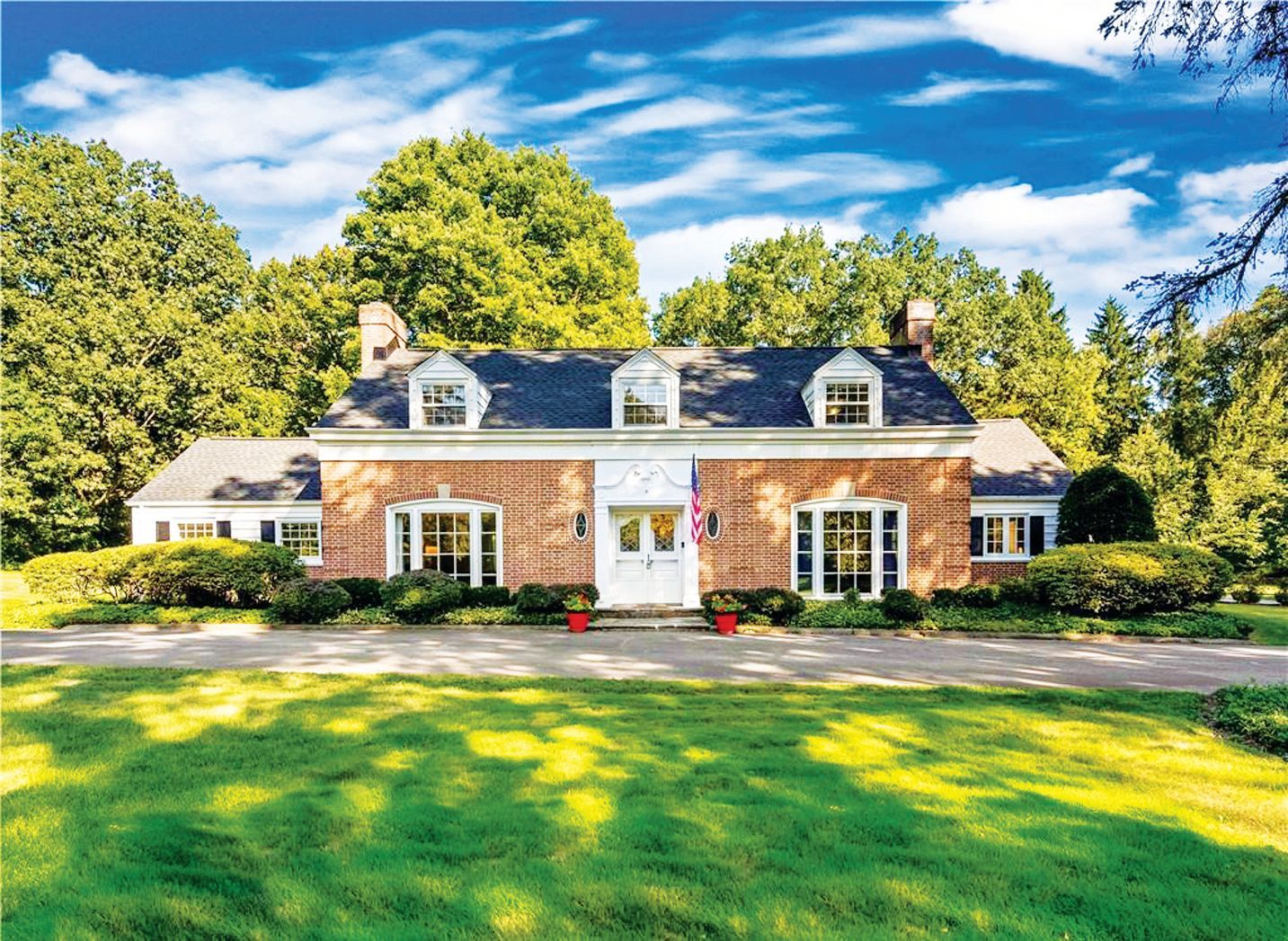A two-story red brick house with white trim, dormer windows, and a black roof, set behind a green lawn and trees.