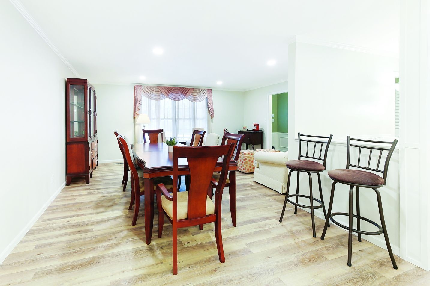 Dining area with a wooden table, six chairs, two bar stools, and a tall display cabinet on a light wood-look floor.