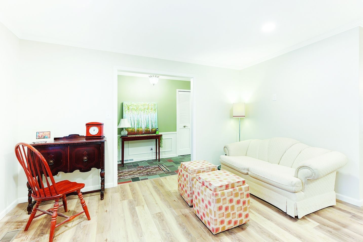 A bright living room features a white sofa, two patterned ottomans, a red wooden chair, and a dark wood side table.