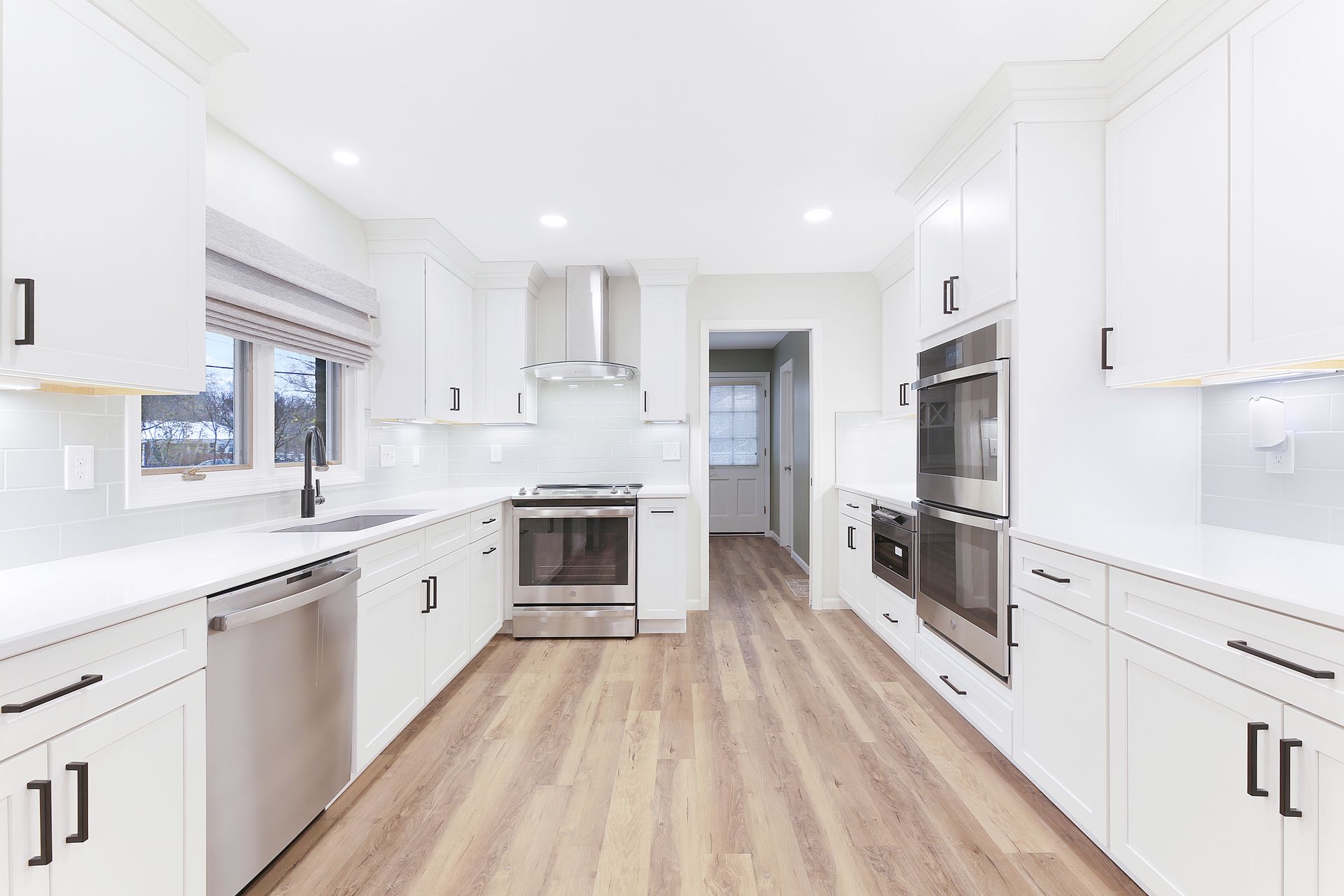 A modern kitchen with white cabinets, stainless steel appliances, white countertops, and light wood-look floors.