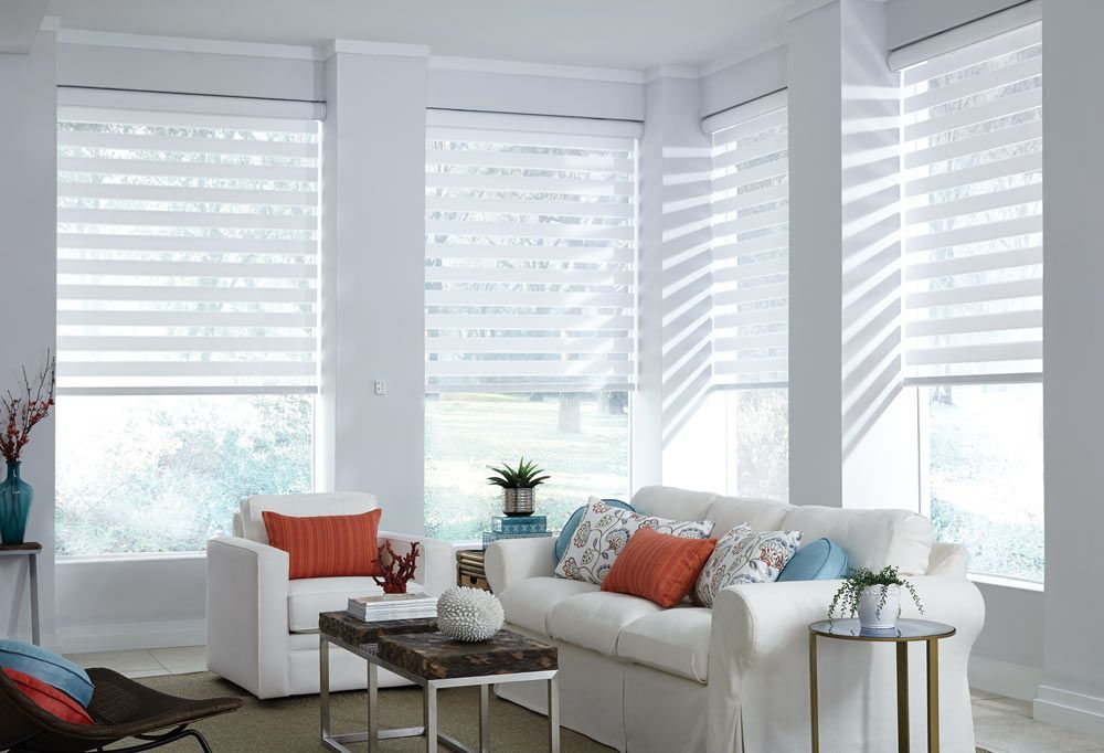Bright living room with white sofas, orange pillows, and striped day-night blinds covering large windows.