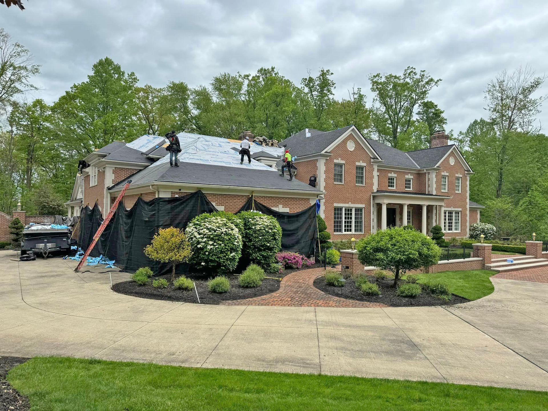 Construction workers install a new dark gray roof on a large brick house surrounded by trees.