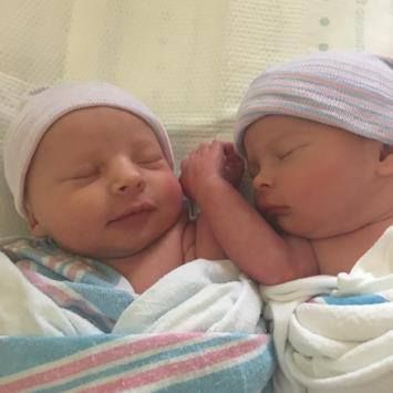 Two swaddled newborns wearing hospital caps, resting side-by-side with their heads touching.