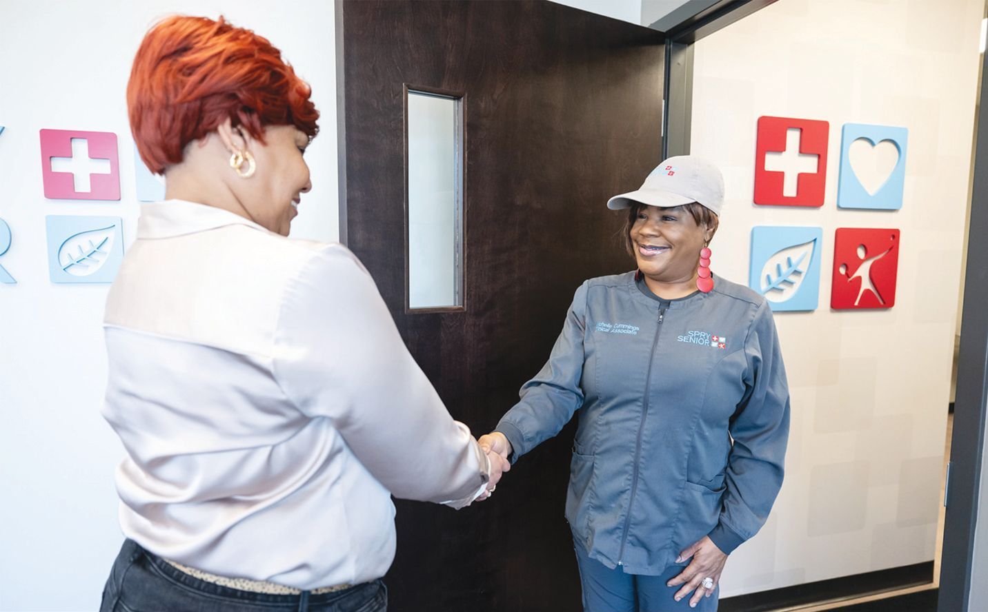 Two people shaking hands in a clinic hallway decorated with colorful medical and wellness icons.