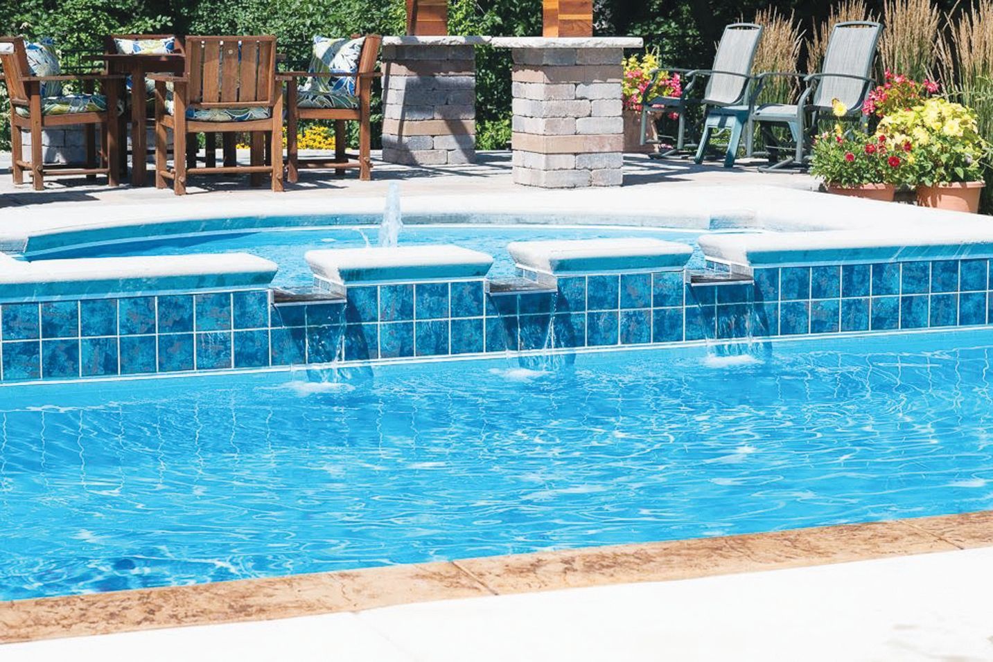 A turquoise swimming pool featuring a tiled spillway with multiple water fountains, set on a sunny patio with deck chairs.