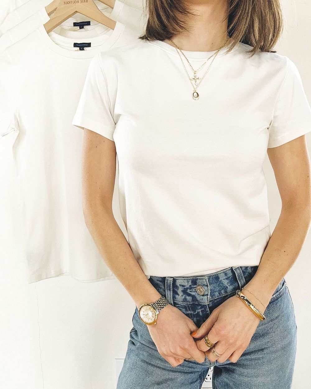 A person wearing a plain white t-shirt, blue jeans, a necklace, and a watch, standing in front of hanging white shirts.