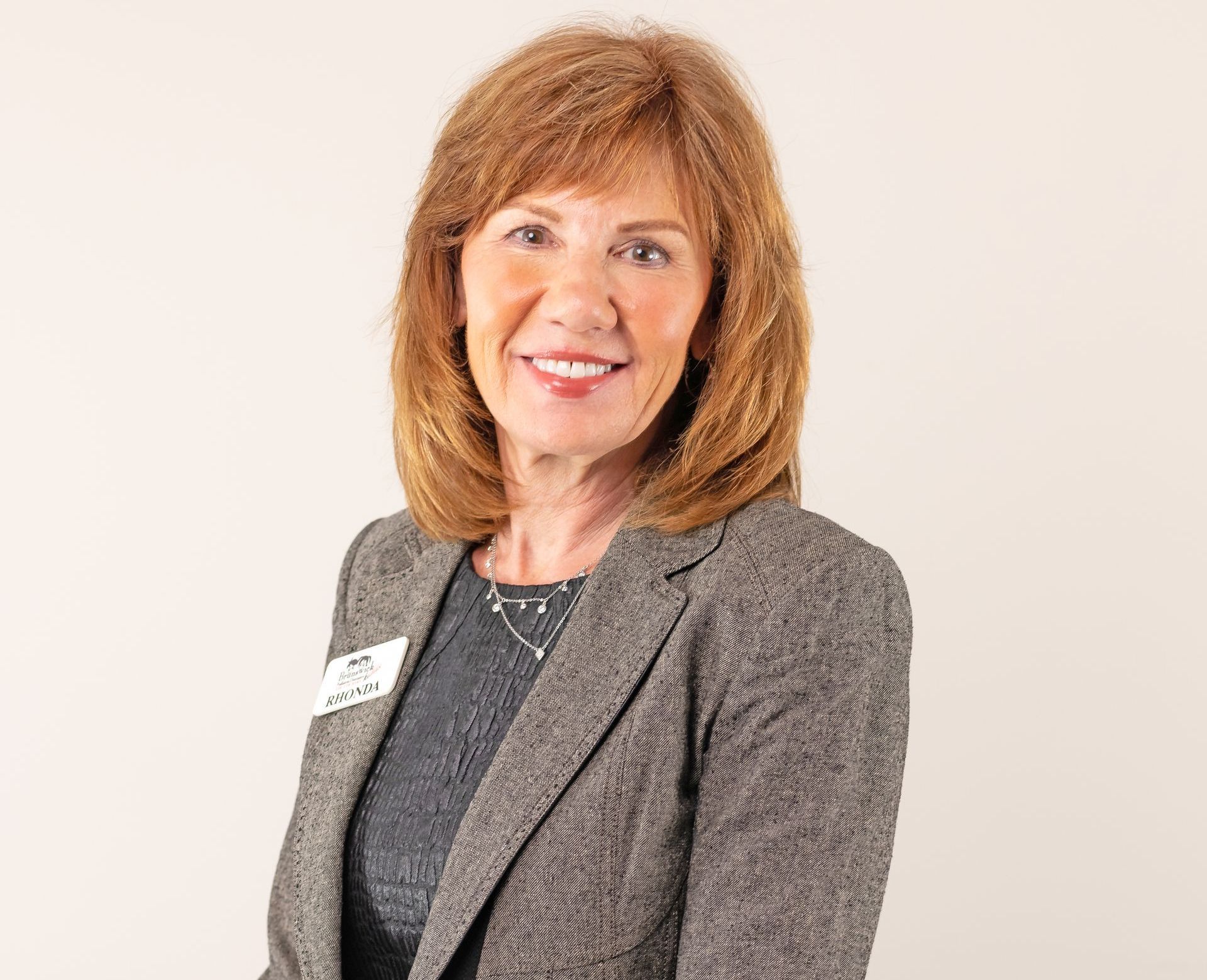 A smiling professional wearing a grey blazer and a small name tag against a neutral background.
