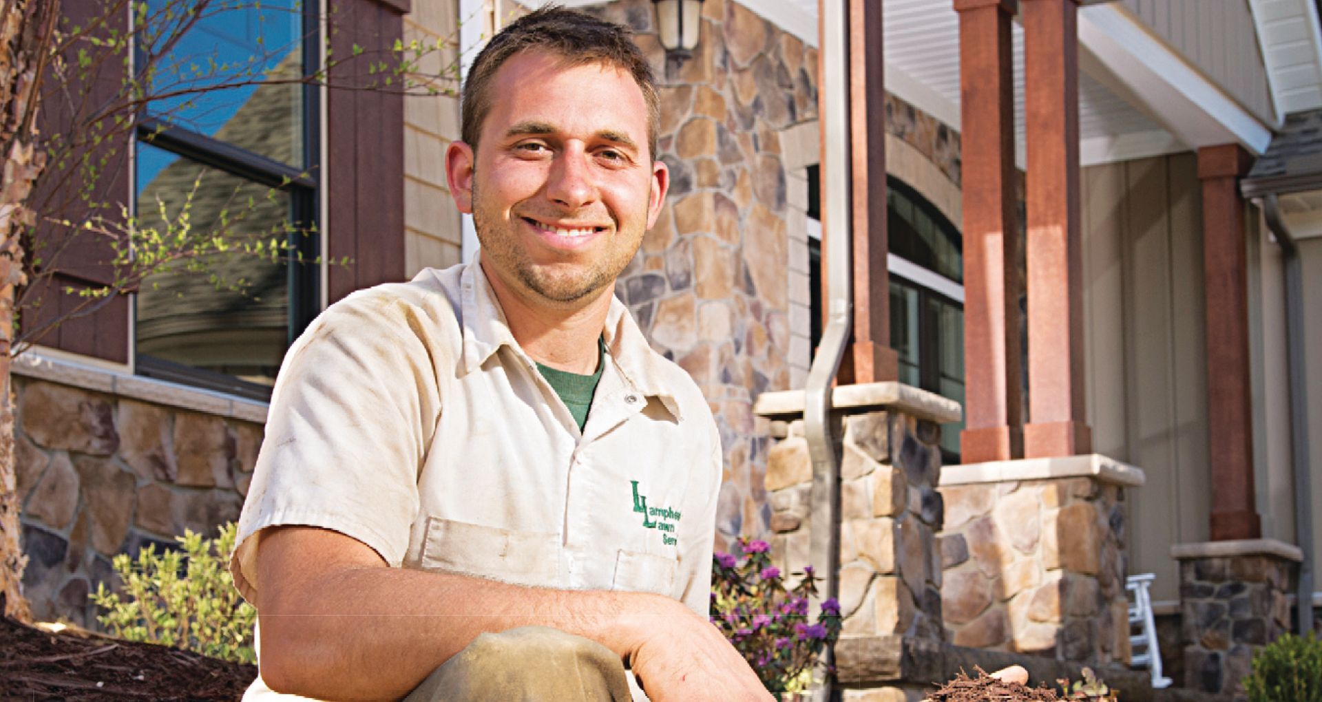 A smiling person in a light-colored work shirt kneels in front of a stone-accented building.