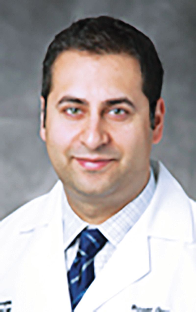 A professional headshot of a person in a white lab coat, light-patterned shirt, and blue tie, smiling against a gray background.