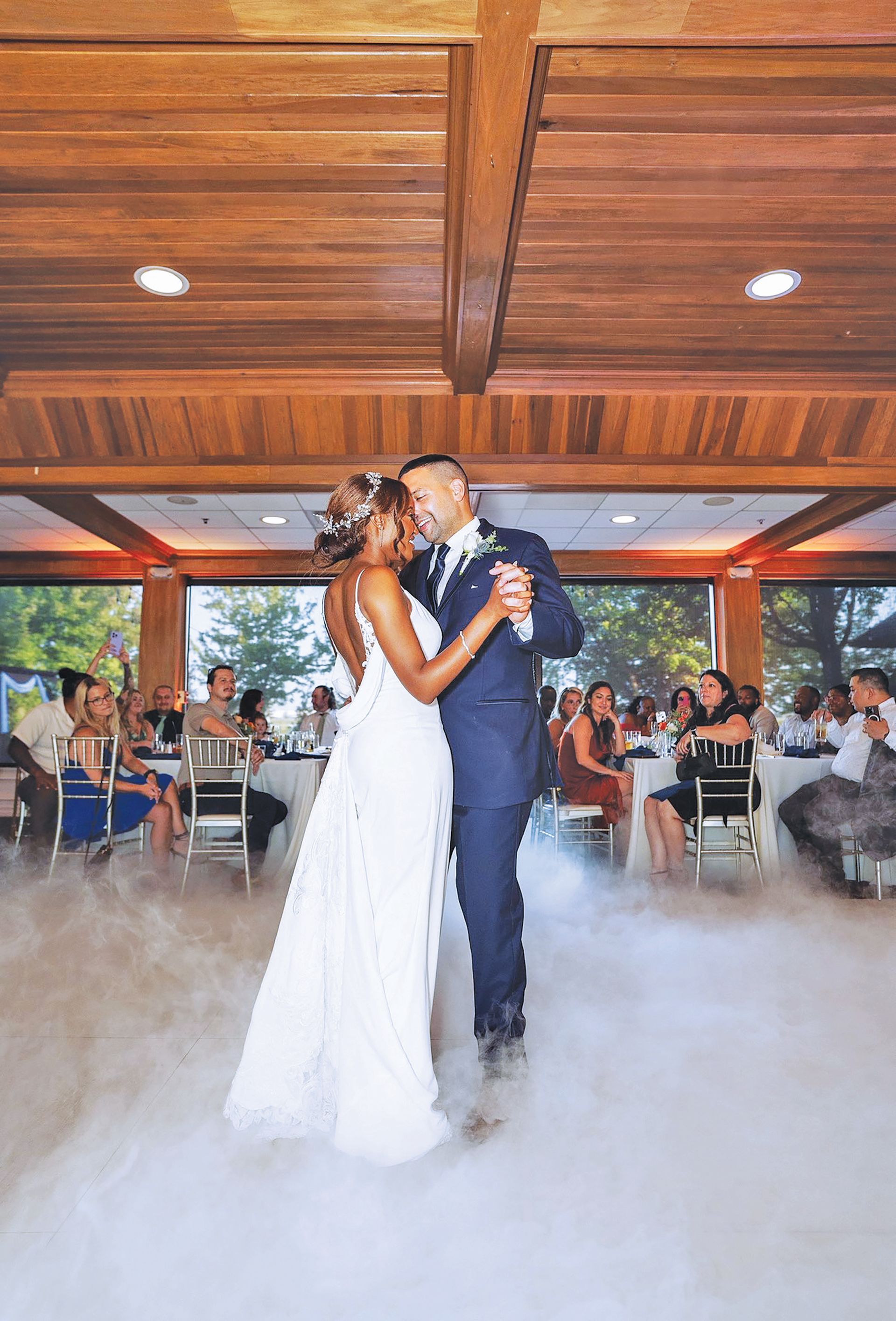 A couple shares a first dance on a floor covered in low-lying fog inside a room with wood-paneled ceilings.