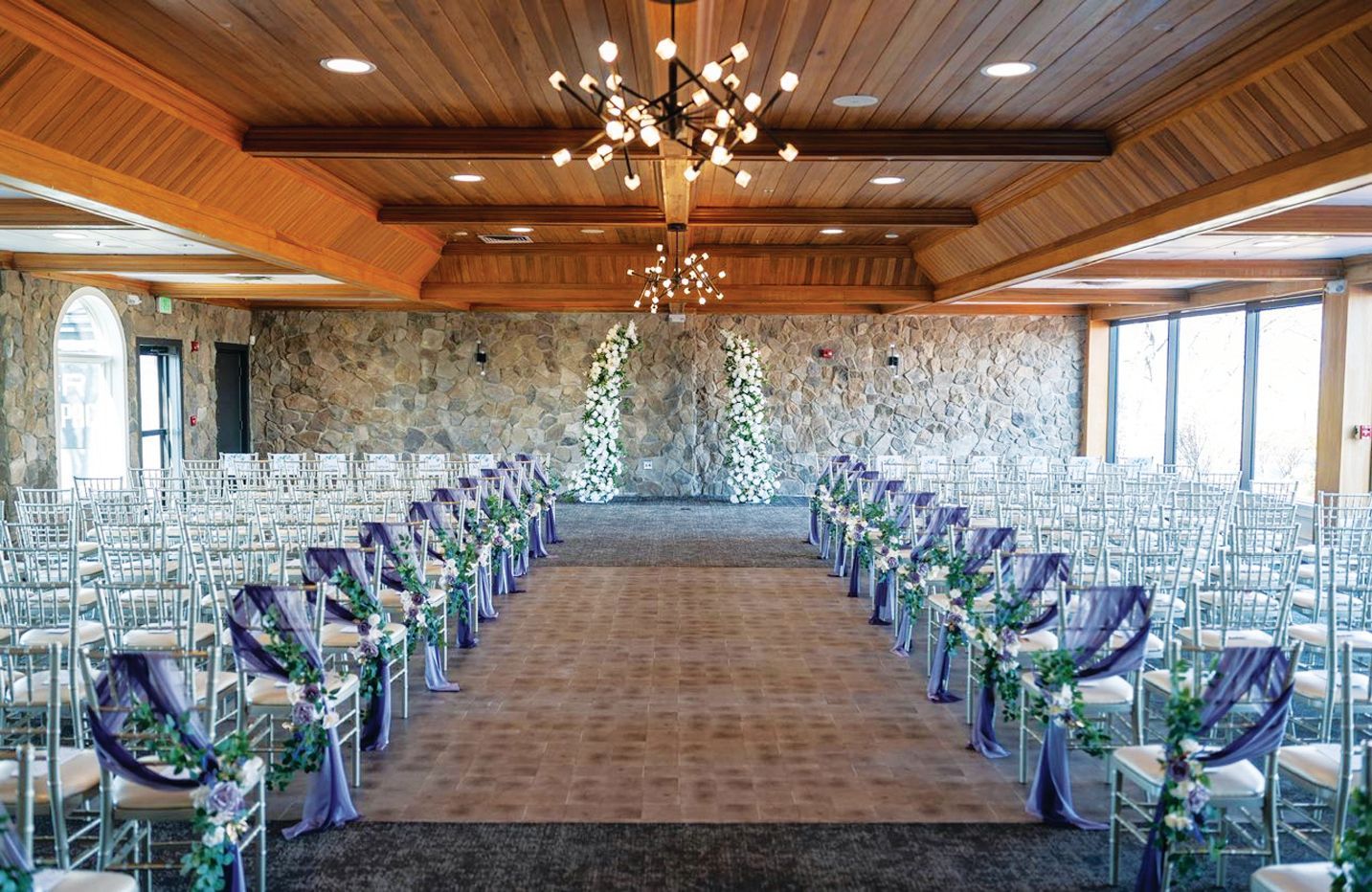 Wedding ceremony setup with rows of chairs decorated with purple sashes and white flowers facing a stone wall with an arch.