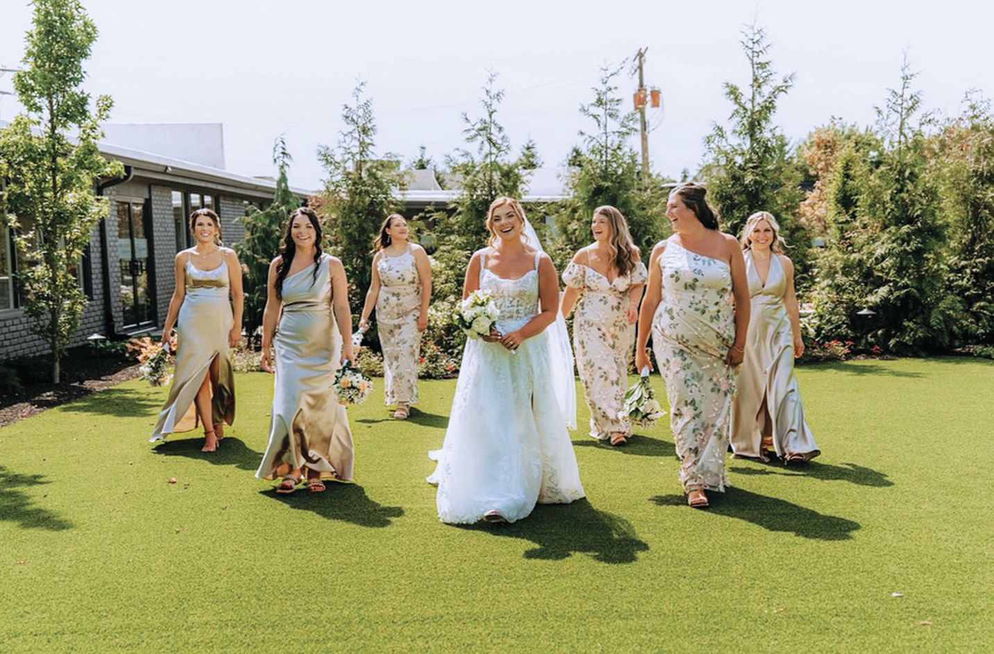 A bride in a white wedding gown walks across a lawn followed by six bridesmaids in flowing, floral-patterned dresses.