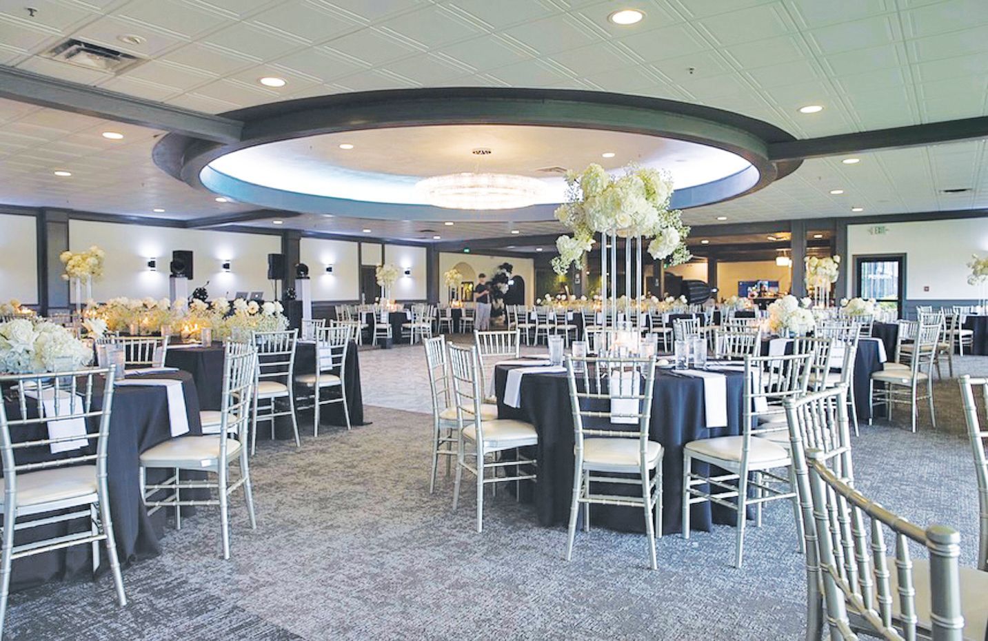 A banquet hall set for a formal event with round tables, dark tablecloths, silver chairs, and white floral centerpieces.