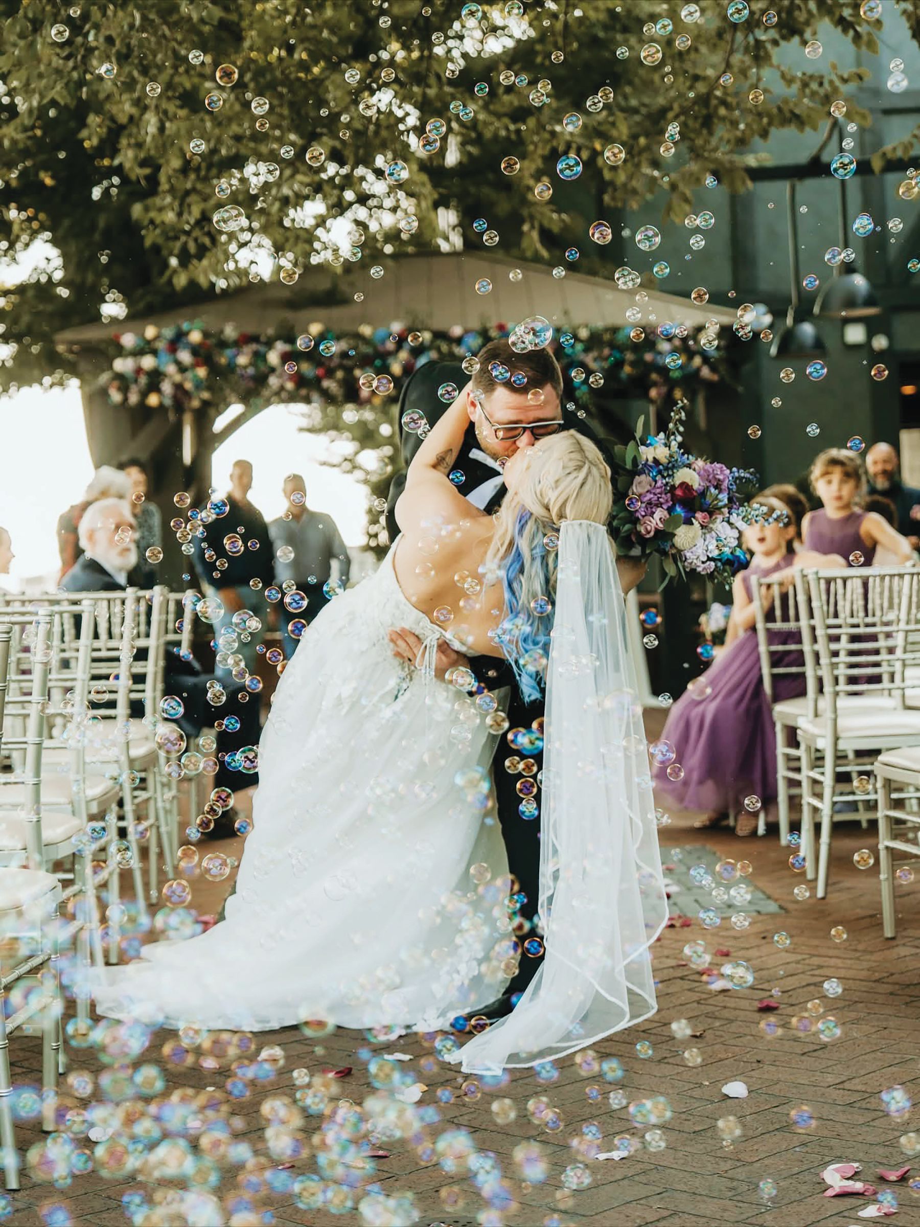 A bride and groom kiss during their wedding ceremony outdoors, surrounded by floating bubbles and guests in chairs.