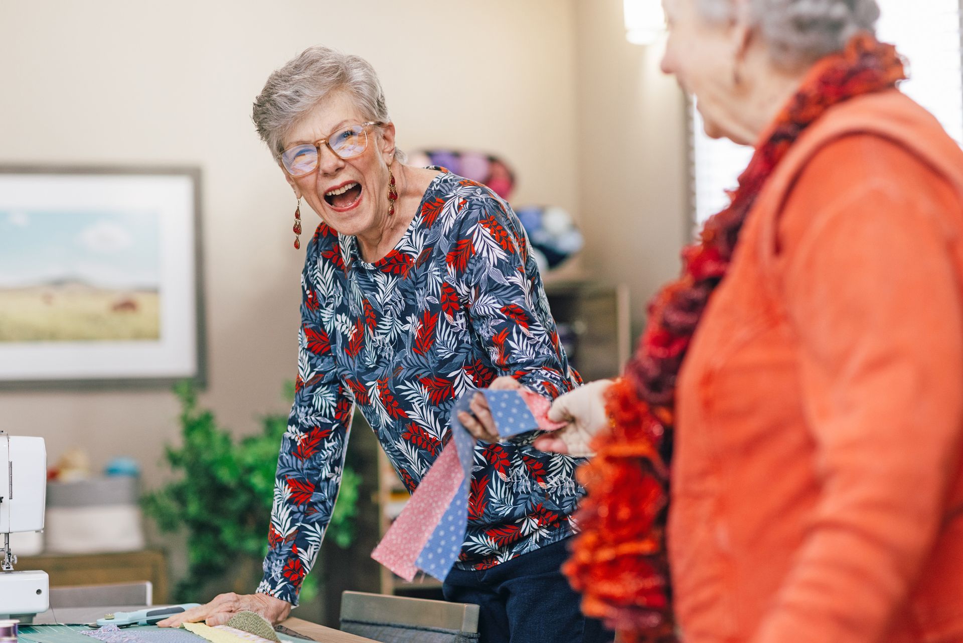 Two people in a craft room share a laugh while one hands a patterned fabric strip to the other.