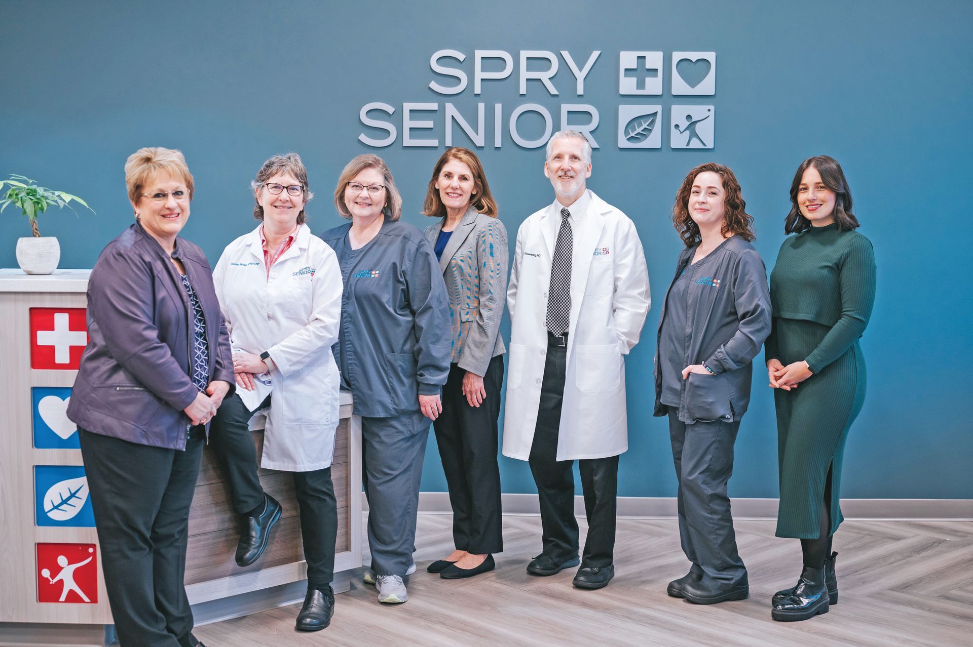 Seven professional staff members stand in front of a blue wall with the SPRY Senior logo and office icons.