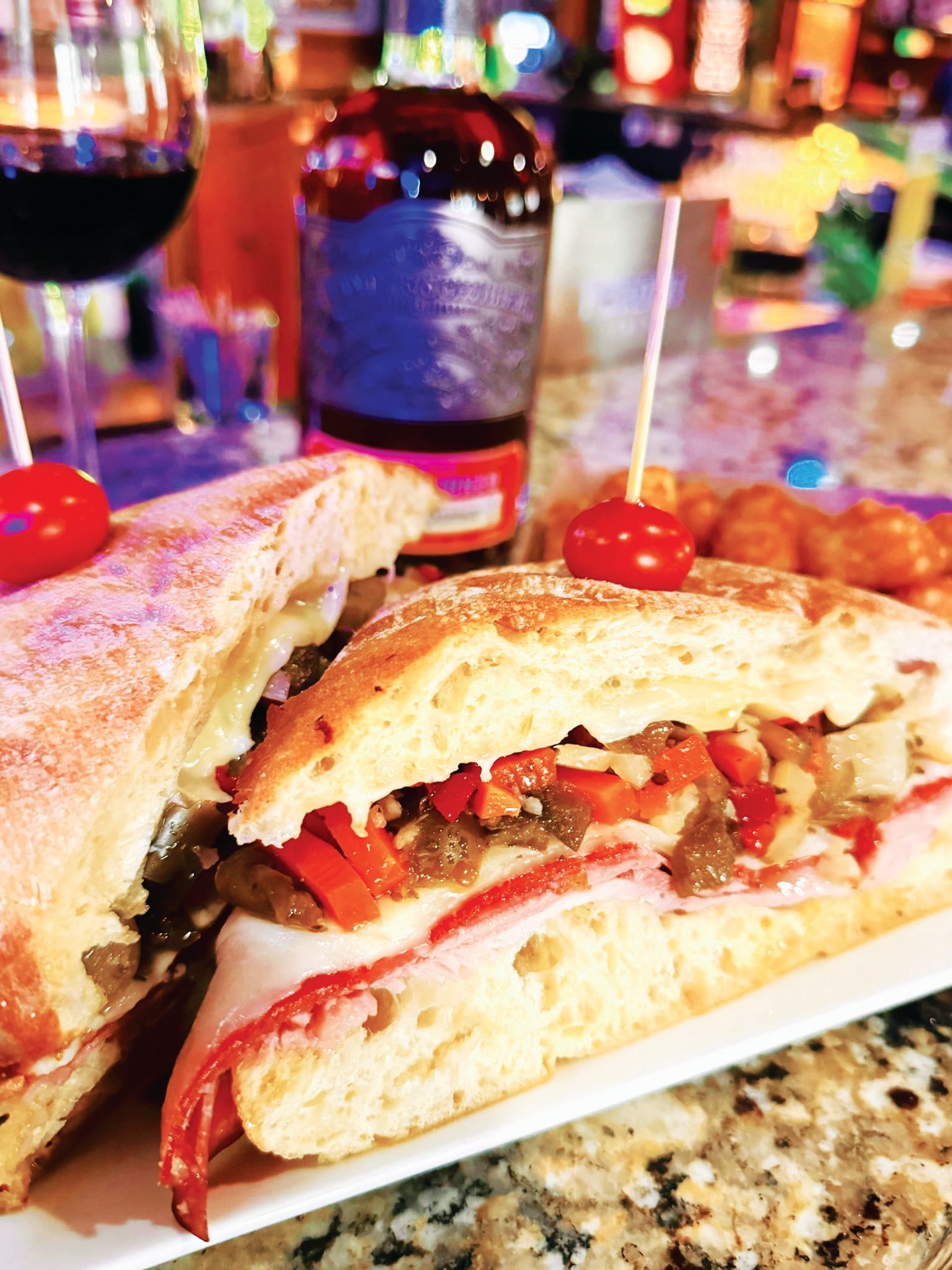 A gourmet panini sandwich with peppers and meat on a plate, with a bottle and a glass of wine in the background.
