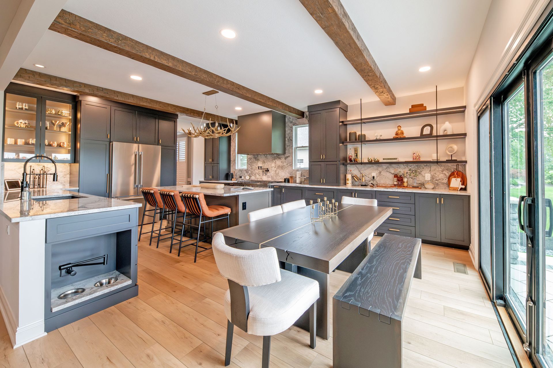 A modern kitchen with dark cabinetry, a central island with bar stools, a long dining table, and light wood flooring.