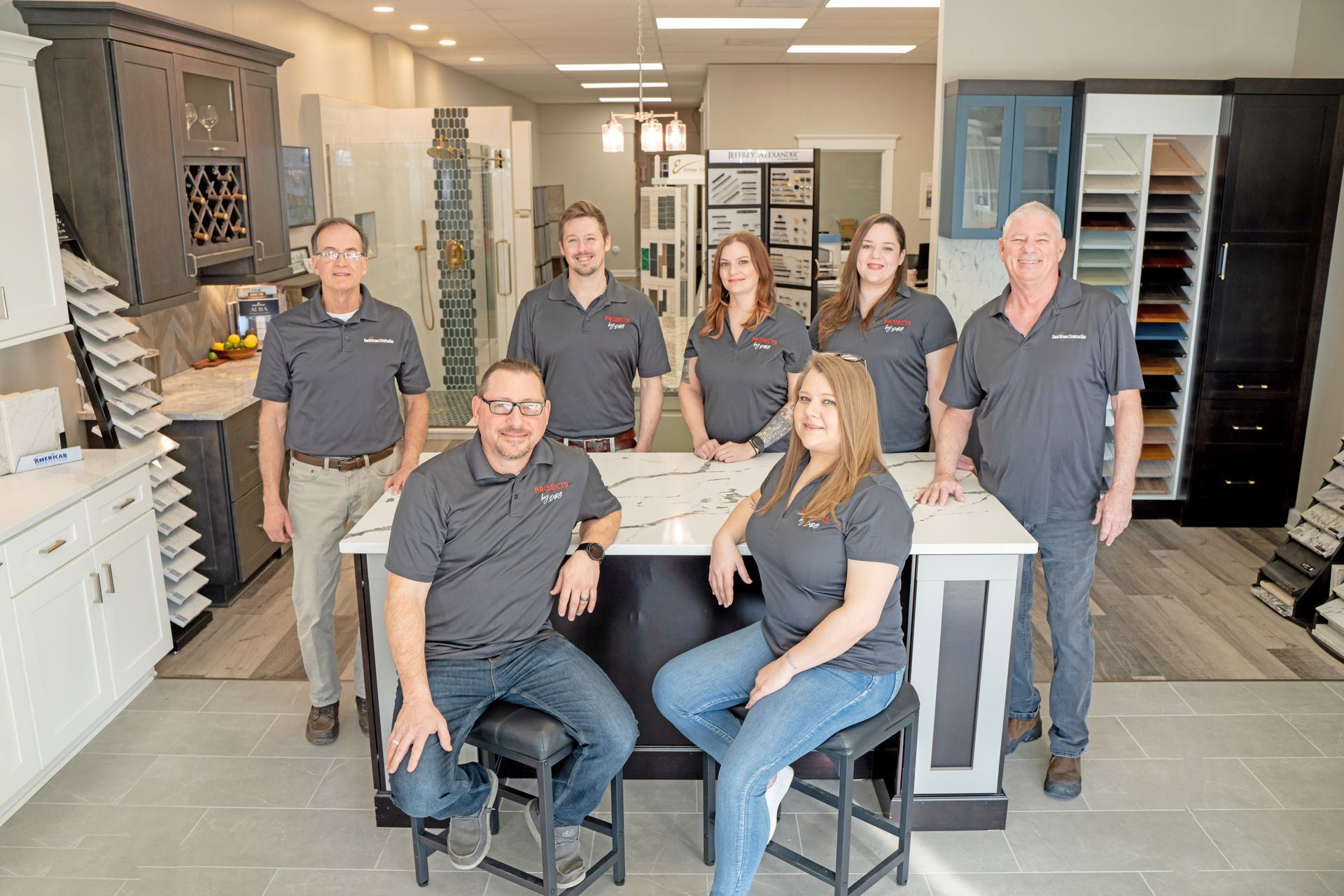 Seven people in matching grey polo shirts pose for a group photo inside a kitchen cabinetry showroom.