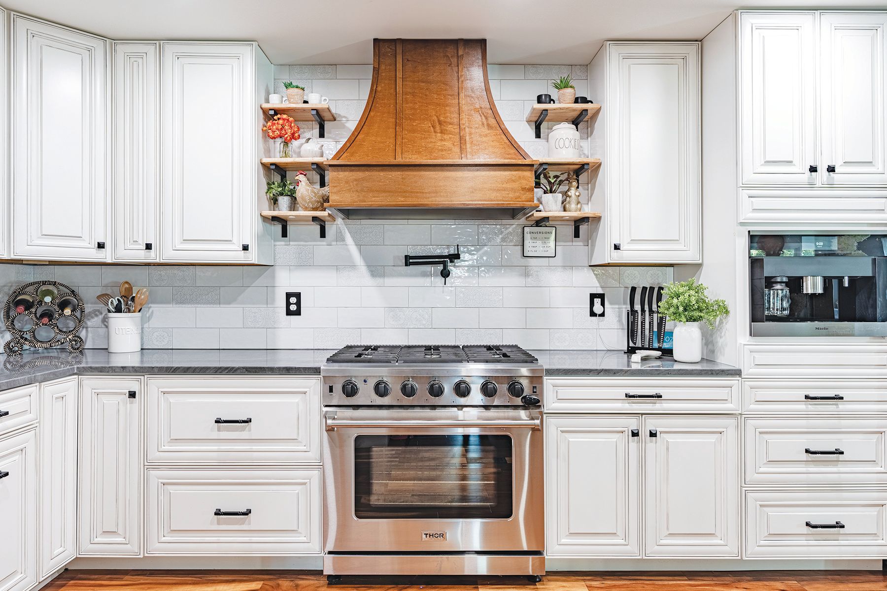 A bright kitchen features white cabinets, a large stainless steel gas stove, and a prominent wood vent hood over the range.