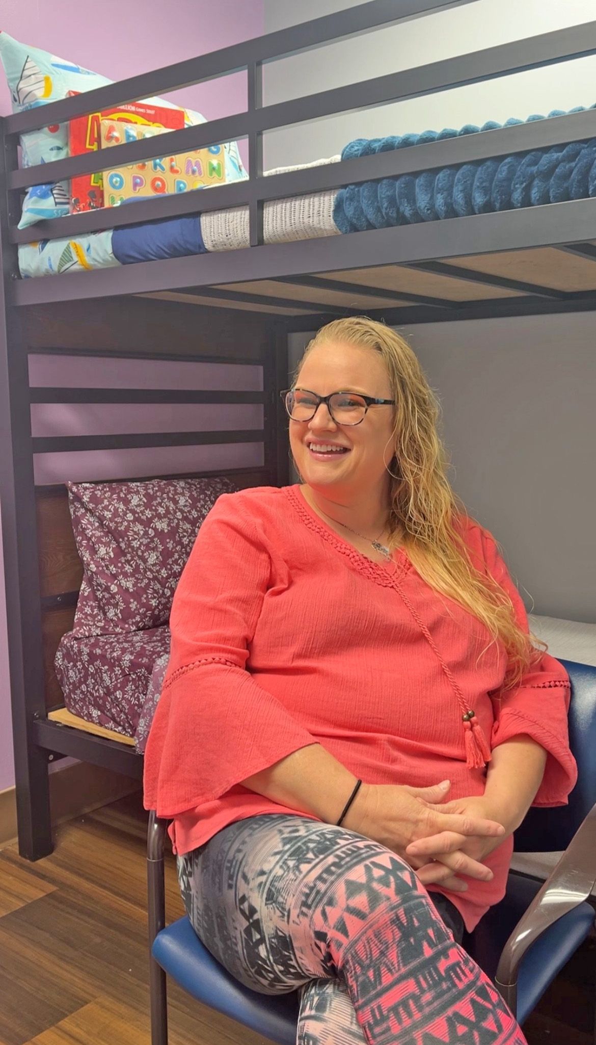 A smiling person wearing a coral-colored top sits in a chair in front of a dark-framed bunk bed.