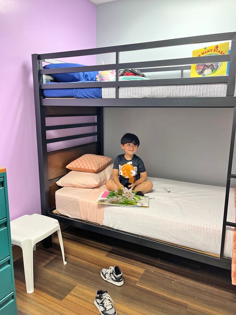 A smiling child sits on the bottom bunk of a dark gray metal bed frame in a room with purple and light blue walls.